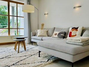 Een moderne woonkamer met een grijze bank, een klein rond tafeltje en een groot raam met gordijnen.