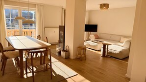 Ein modernes Wohnzimmer mit Sofa, Fernseher, Esstisch mit Stühlen und einem Fenster mit Vorhängen.