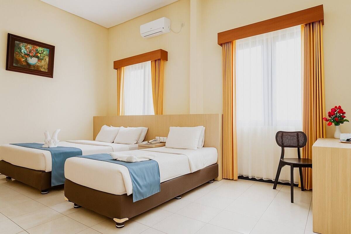 Une chambre d’hôtel avec deux lits, un bureau et une chaise. Il y a un tableau au mur et un appareil de climatisation au-dessus des lits.