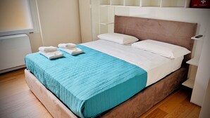 Una cama bien hecha con una colcha verde azulada y dos toallas blancas dobladas, piso de madera y una mesita de noche blanca con un botiquín rojo de emergencia.