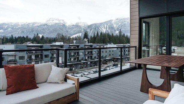 En balkon med et træbord og sofa, der udover en moderne bygning og sneklædte bjerge.