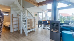 A spacious living area with a wooden staircase, a dining table with chairs, and a large window providing a view of the outside. - Coastal Family Escape with Enclosed Garden (Renesse)