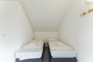 A small, single-room hotel room with two single beds, a wooden bench, a wall-mounted light fixture, and a power outlet. - Coastal Family Escape with Enclosed Garden (Renesse)