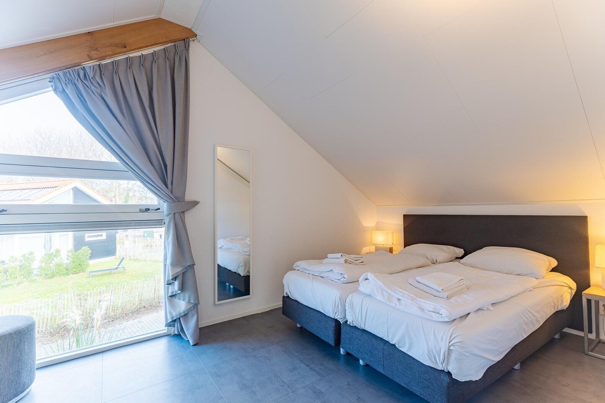 Un dormitorio con cama, lámpara de noche, ventana con cortinas y vistas al exterior.