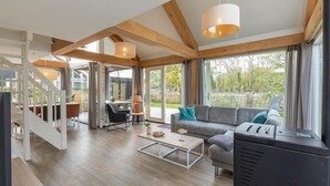 A spacious living area with a sofa, coffee table, and a television. There is a staircase leading to an upper level, and large windows providing a view of the outdoors. - Zeeland Luxury Villa near Beach (Renesse)