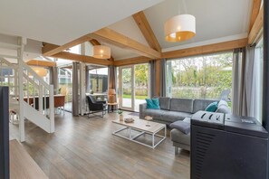 Ein geräumiger Wohnbereich mit einer Couch, einem Couchtisch und einem Fernseher. Eine Treppe führt zu einer oberen Ebene, und große Fenster bieten einen Blick ins Freie.