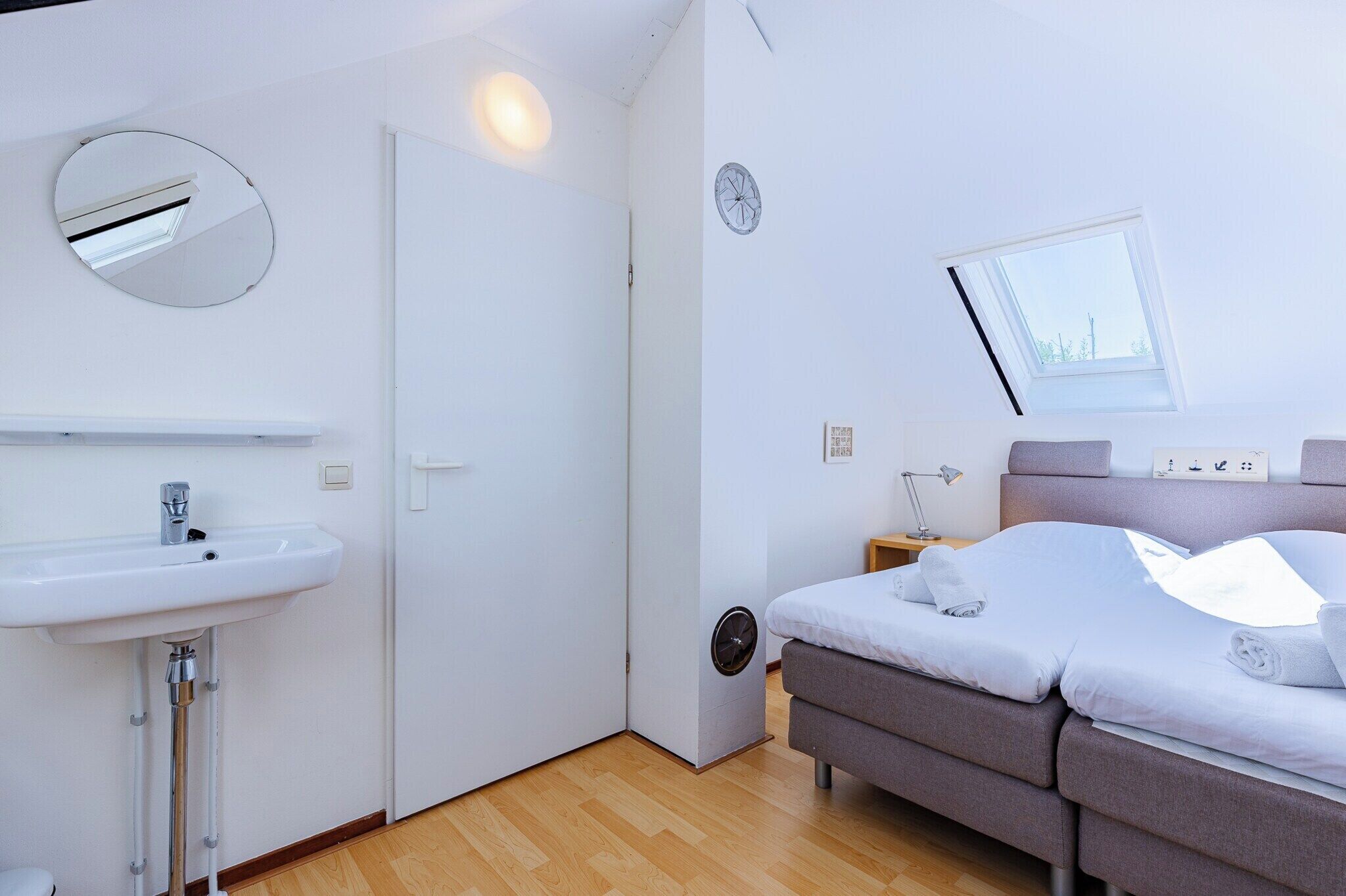 Une chambre à coucher moderne avec un lit, une table de chevet, une lampe, un miroir rond, une fenêtre et un lavabo blanc.