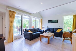 Un salon spacieux avec deux canapés noirs, une table basse en bois et un fauteuil en osier. De grandes fenêtres, ornées de rideaux, laissent entrer la lumière naturelle et offrent une vue sur la verdure environnante.