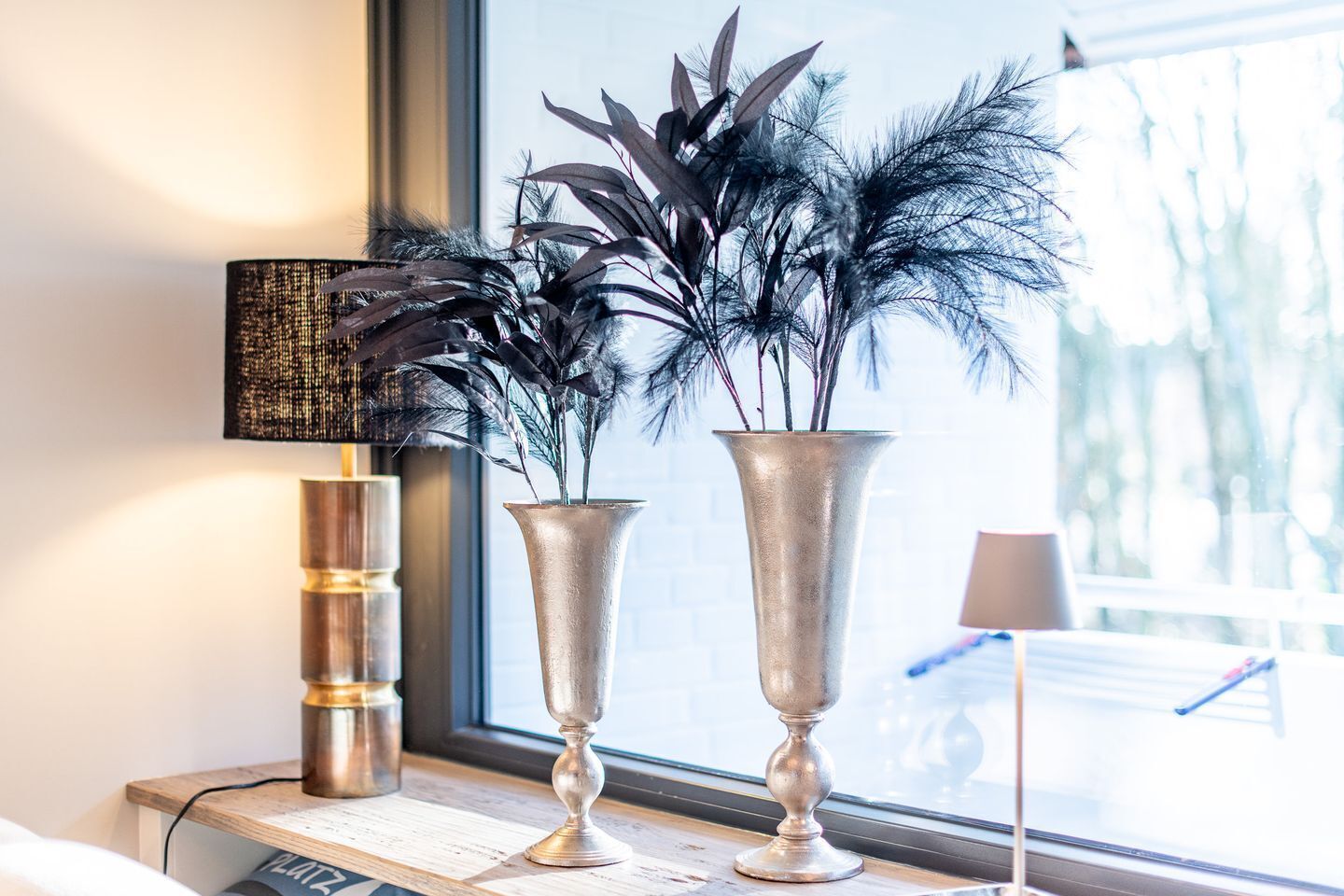A window sill with two silver vases containing plants, a lamp, and a laptop.