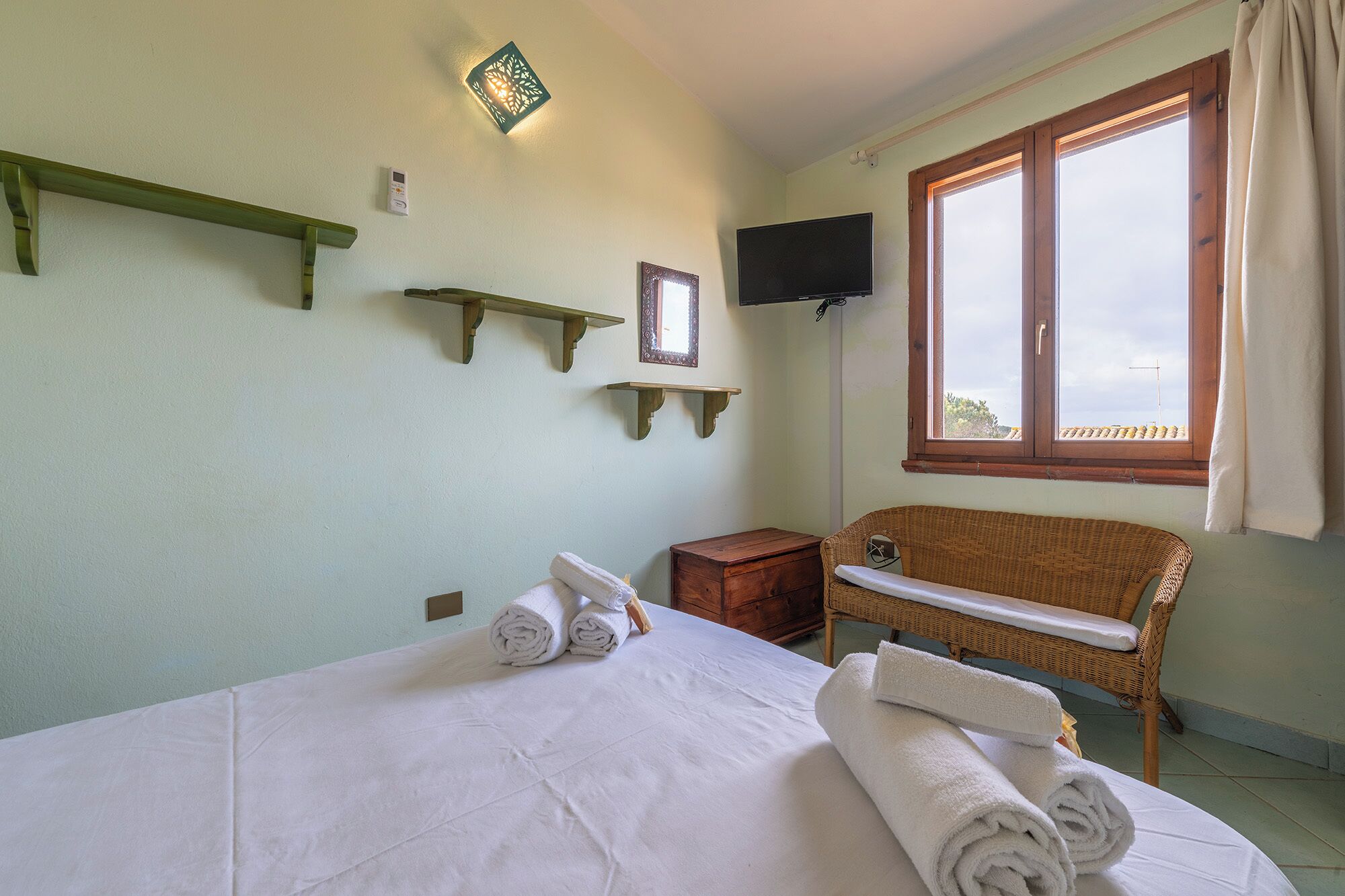Ein Hotelzimmer mit einem Bett, einem Nachttisch aus Holz, einem Korbstuhl, einem an der Wand montierten Fernseher und einem Fenster mit Vorhängen.