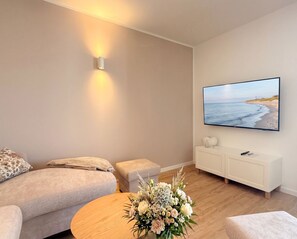 A living room with a sofa, a coffee table with flowers, a TV on the wall, and a small cabinet. - 2170 - Gode Ström quarters "Söven" - Penthouse Strandwiese - 2170 - Gode Ström (Dahme)