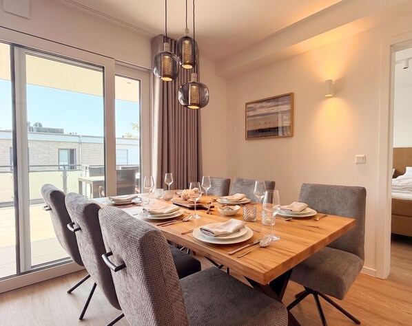 A dining area with a wooden table, grey upholstered chairs, and a view through a glass door. - 2170 - Gode Ström quarters "Söven" - Penthouse Strandwiese - 2170 - Gode Ström (Dahme)
