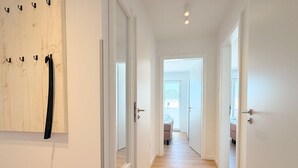 Un couloir doté d’un plancher en bois, de murs blancs et d’une porte donnant sur une pièce où se trouvent un lit et une fenêtre.