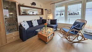 A living room with a sofa, a glass coffee table, a wicker chair, a mirror, and a view of the city through large windows. - Vacation apartment Lüttje Mööv Norddeich - Comfortable vacation apartment Lüttje Mööv (Norden)