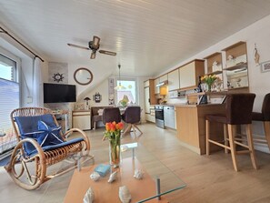 A modern kitchen with wooden cabinets, a dining area with wicker chairs, and a living room with a large window. - Vacation apartment Lüttje Mööv Norddeich - Comfortable vacation apartment Lüttje Mööv (Norden)