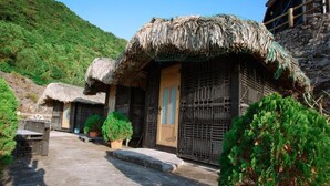 A traditional thatched-roof house with a wooden door and window grills, surrounded by greenery and a stone wall. - Cat Ba Mountain View Homestay (Hai Phong)