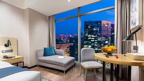 Camera d'albergo con un'ampia finestra che offre vista sulla città di notte, un divano, una poltrona, un piccolo tavolo con della frutta e un comodino con una lampada.