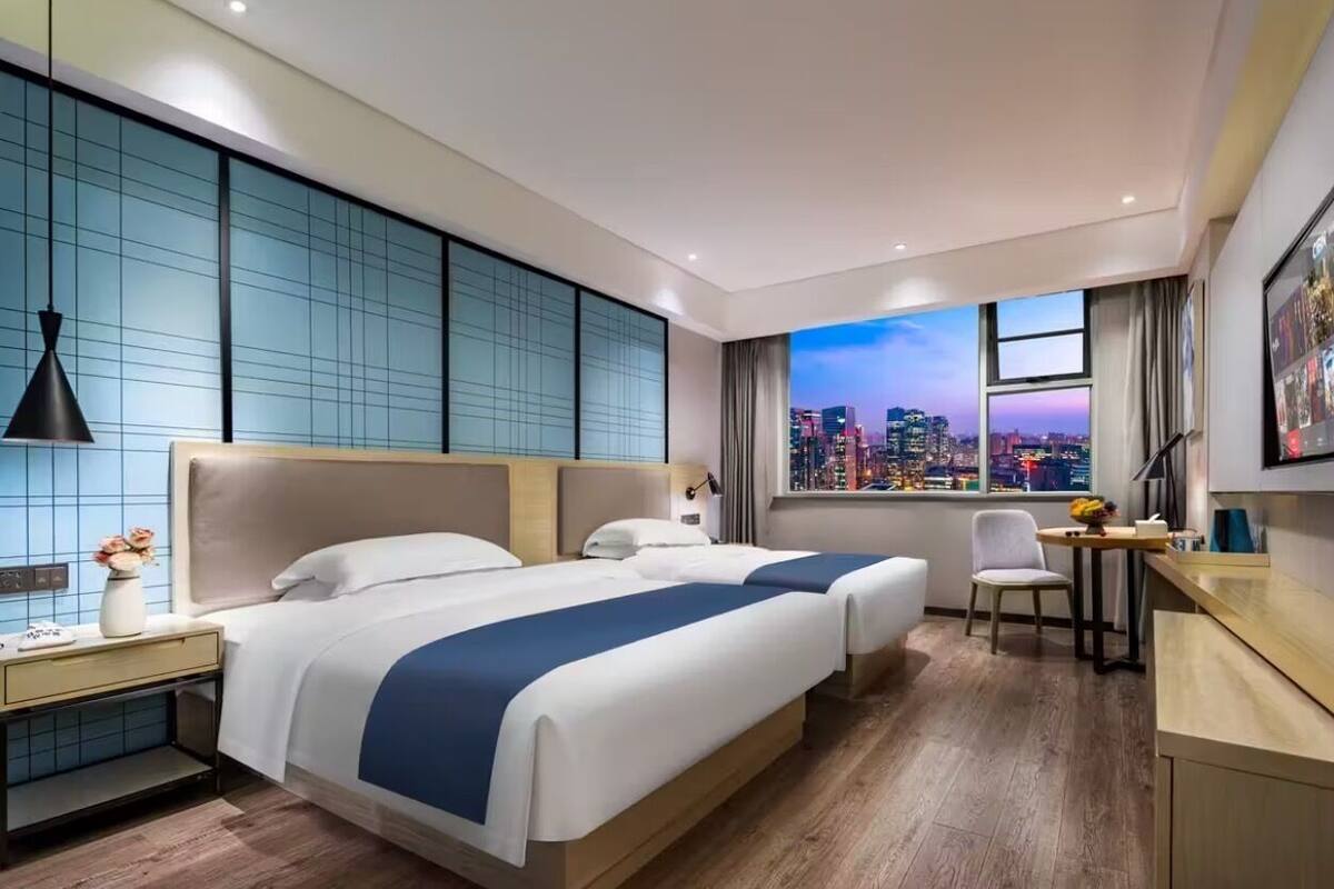A modern hotel room with a city view, two beds, a desk, and a chair.