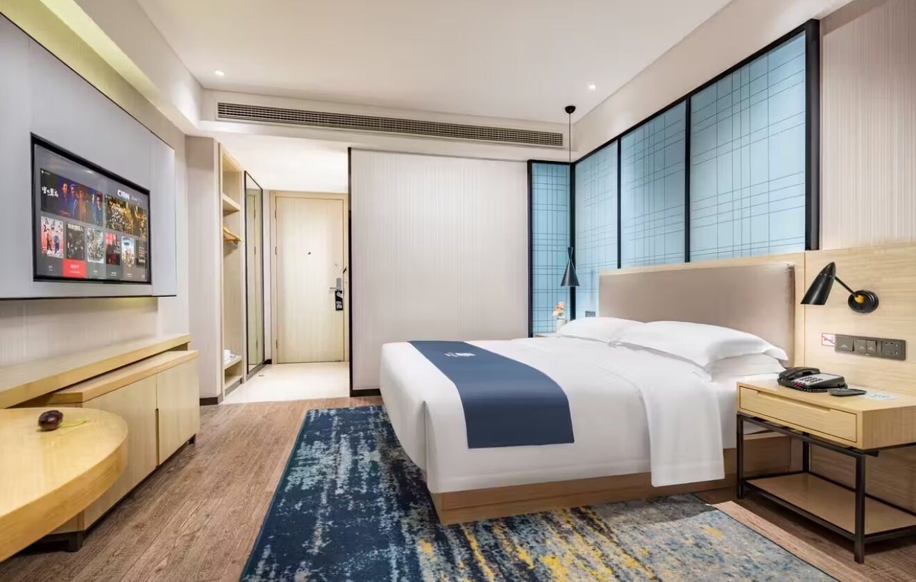A modern hotel room with a large bed, a desk, a TV mounted on the wall, and a sliding door leading to a bathroom.
