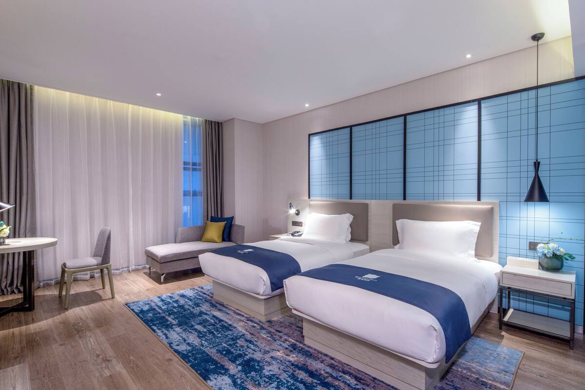A modern hotel room with two beds, a desk, a chair, and a large window with curtains.