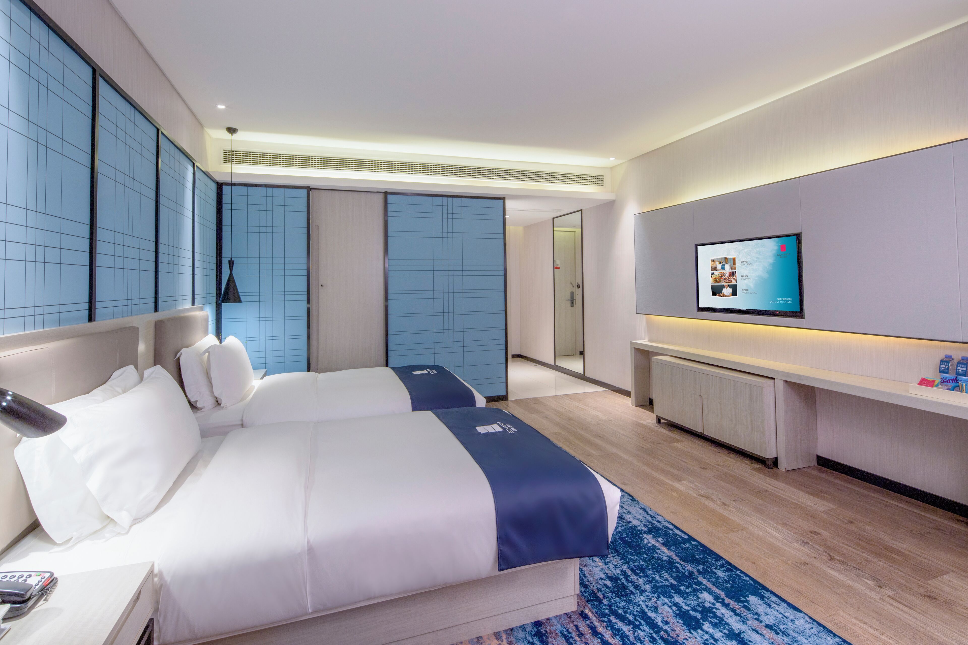 A modern hotel room with two beds, a TV, a desk, and a bathroom visible through a sliding door.