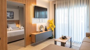 Et moderne hotelværelse med en seng, et tv ophængt på væggen, et konsolbord med dekorative genstande og et stort vindue med lette gardiner.