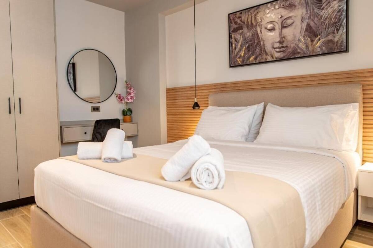 Un lit bien fait, avec du linge de lit blanc et des serviettes pliées, un miroir rond, un tableau représentant Bouddha et une tête de lit en bois.