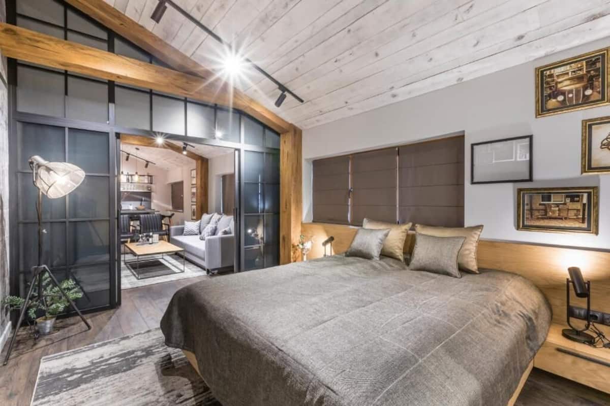 A modern bedroom with a large bed, wooden beams, a desk, and a view into the living area.