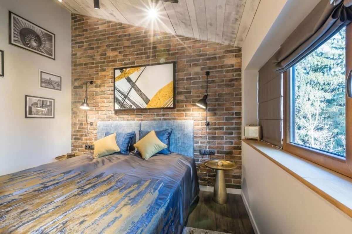 A bedroom with a brick wall, a large bed with blue bedding, and a window with a view of trees.