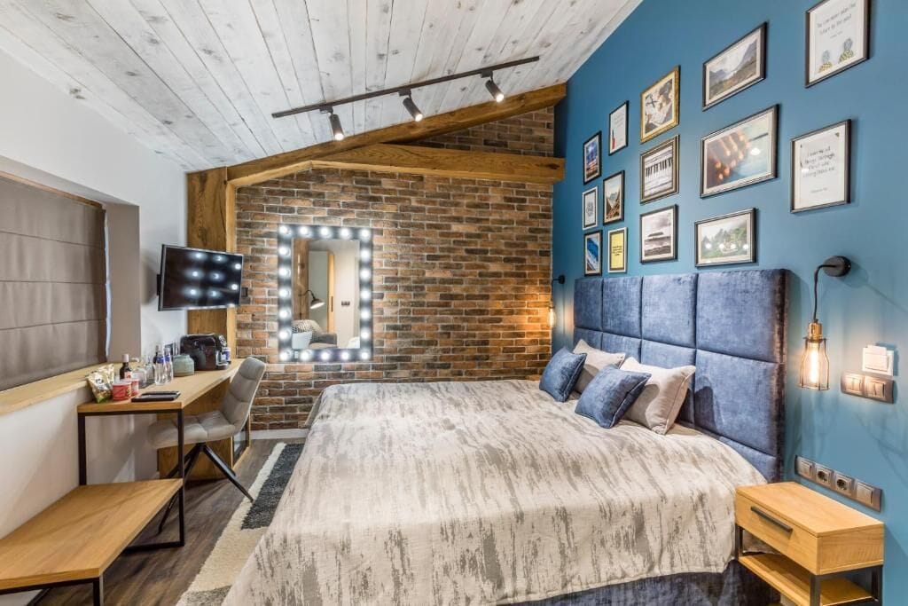 A bedroom with a large bed, a desk, a chair, and a TV. There are framed pictures on the wall and a brick wall behind the bed.