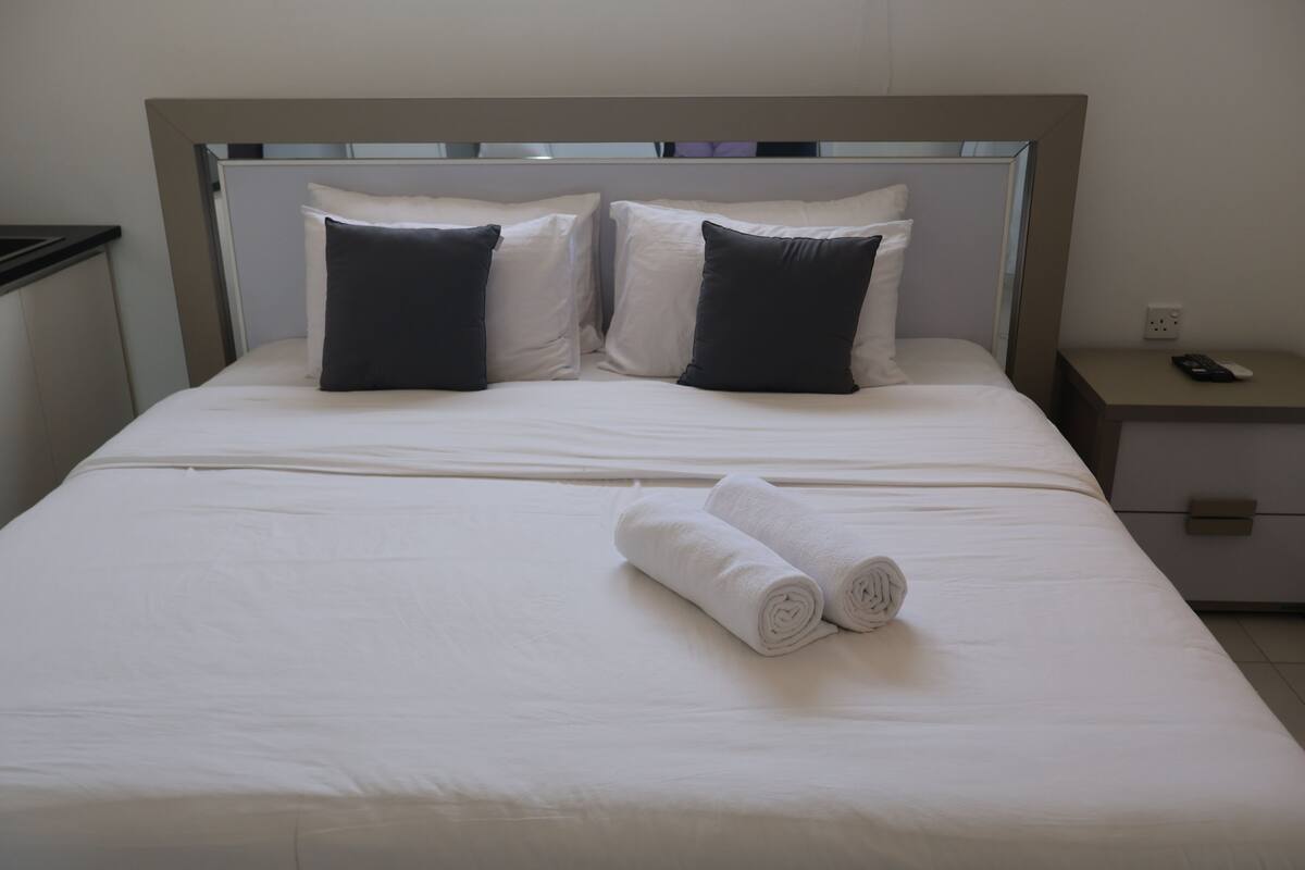 A neatly made bed with white linens and two dark pillows, a bedside table with a remote control, and a power outlet on the wall.