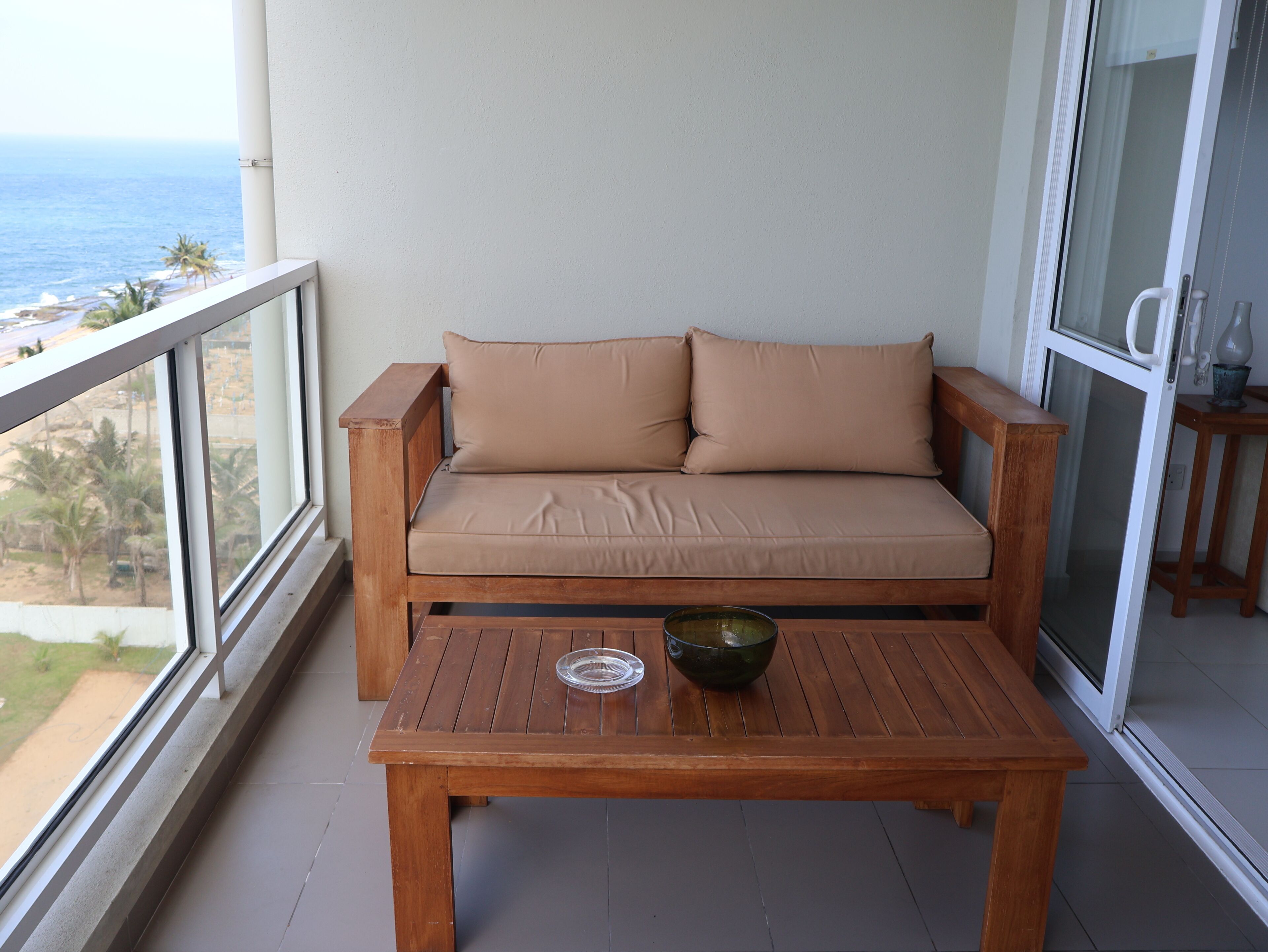 A balcony with a wooden sofa and a table, overlooking a beach and palm trees.