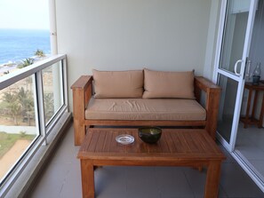 A balcony with a wooden sofa and a table, overlooking a beach and palm trees. - Beach Front 2 by A&A Villas (Uswetakeiyawa)
