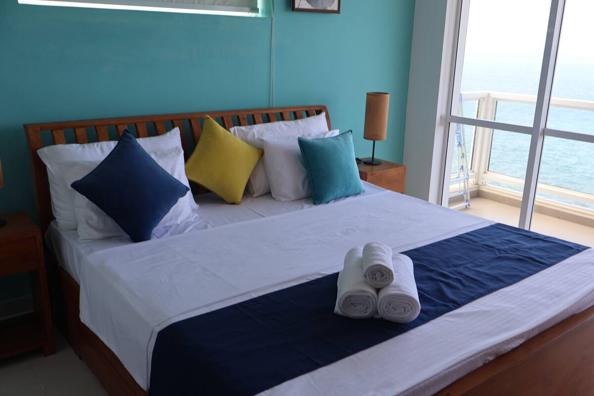 A neatly made bed with white and navy bedding, multiple pillows in various colors, and a set of towels on the bed.
