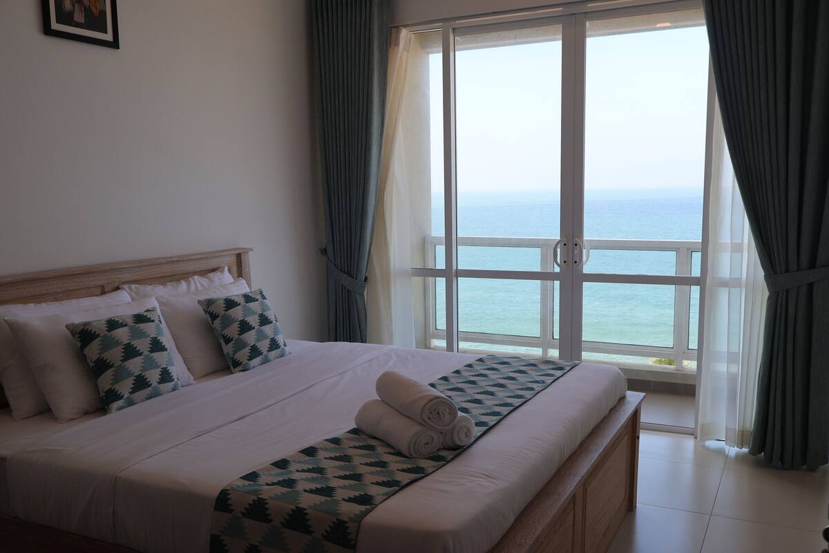 A neatly made bed with a patterned bedspread and pillows, a wooden headboard, and a view of the sea through large windows.