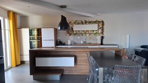 Eine moderne Küche mit Kochinsel, Essbereich mit transparenten Stühlen und einem großen Fenster mit gelben Vorhängen.