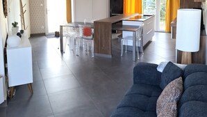 Ein modernes Wohnzimmer mit einem dunklen Sofa, einem Essbereich mit transparenten Stühlen, einer Küche mit schwarzem Kühlschrank und einem großen Fenster mit Vorhängen.