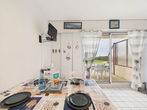 En matplats med ett bord för fyra personer, en tv som är monterad på väggen och utsikt över utomhusmiljön genom en glasdörr.