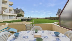 An outdoor patio with a table set for dining, surrounded by white chairs, and overlooking a grassy area with a building in the background. - Apartment with sea view, 2 adults and 2 children (Saint-Gilles-Croix-de-Vie)
