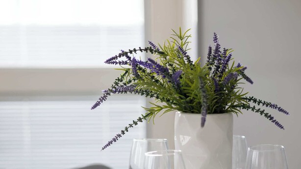 Ein Esstisch, gedeckt mit weißen Tellern, Besteck und klaren Weingläsern, mit einer weißen Vase voller lila Blumen in der Mitte.
