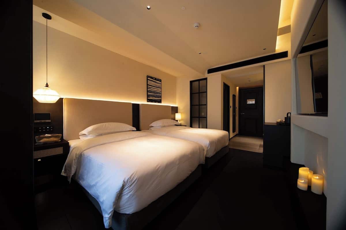 A hotel room with two beds, bedside tables, a hanging lamp, and a view of the hallway.