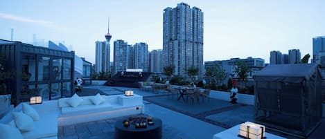 A rooftop terrace with seating areas, a table, and a view of the city skyline including a tall building with a distinctive spire.