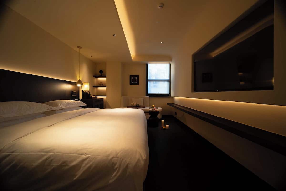 A hotel room with a large bed, bedside tables, a TV mounted on the wall, and a window providing natural light.