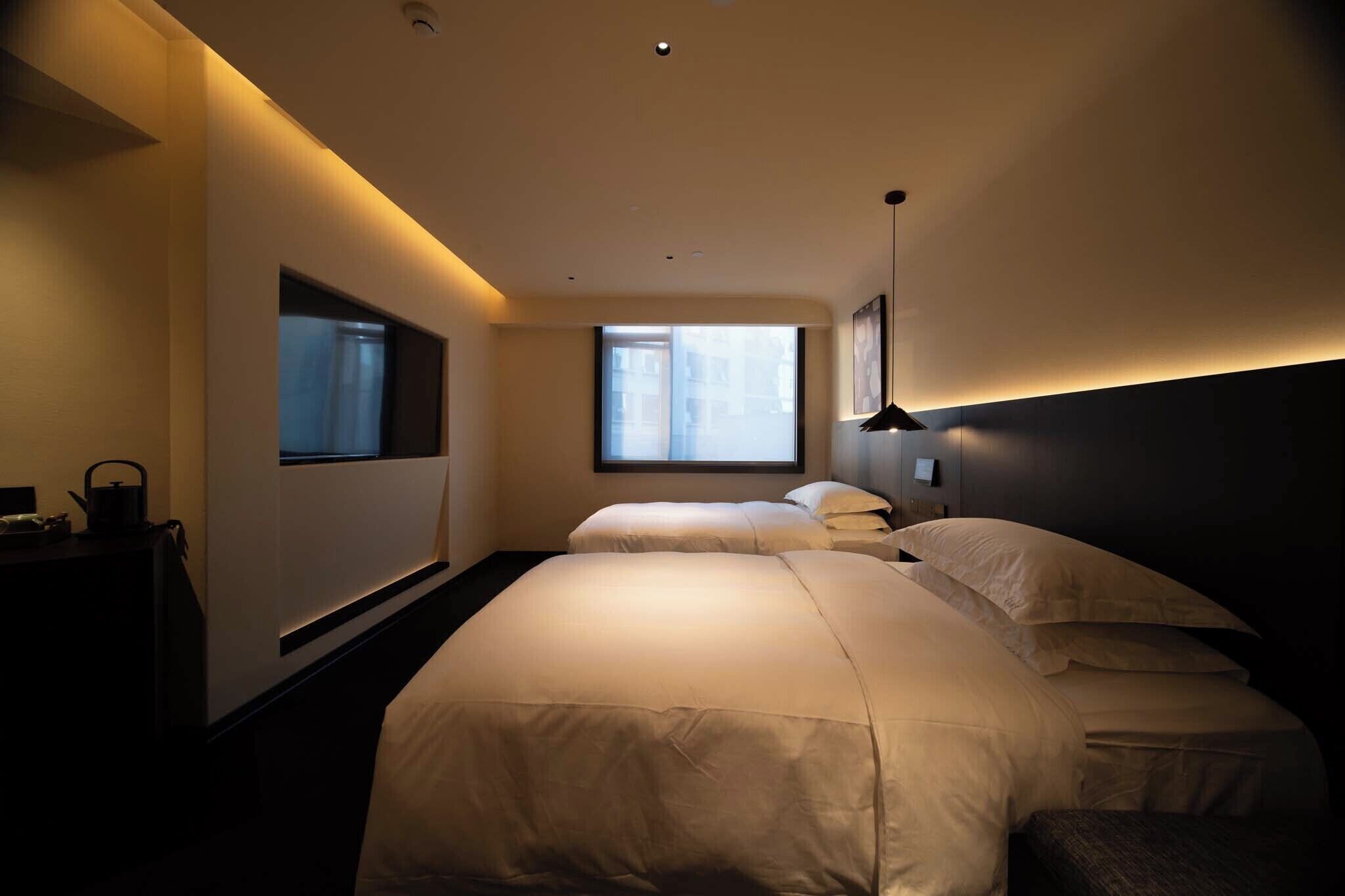 A modern hotel room with two beds, a large window, and a bedside table with a kettle.