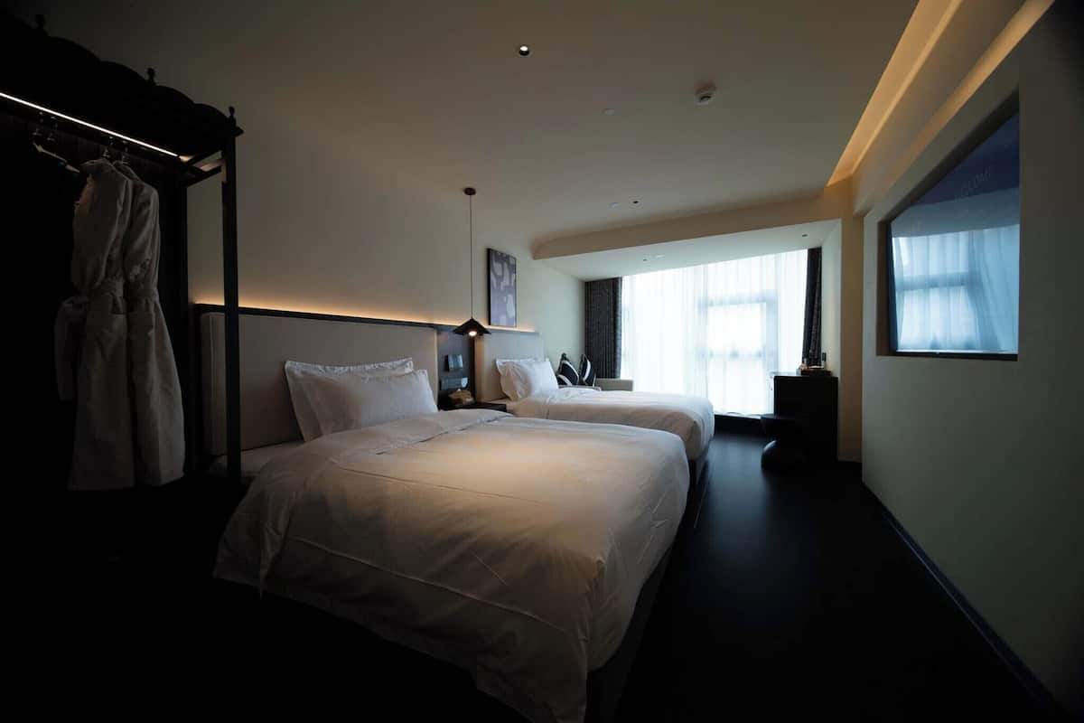 A hotel room with two beds, a robe rack, a bedside table, and a large window with curtains.