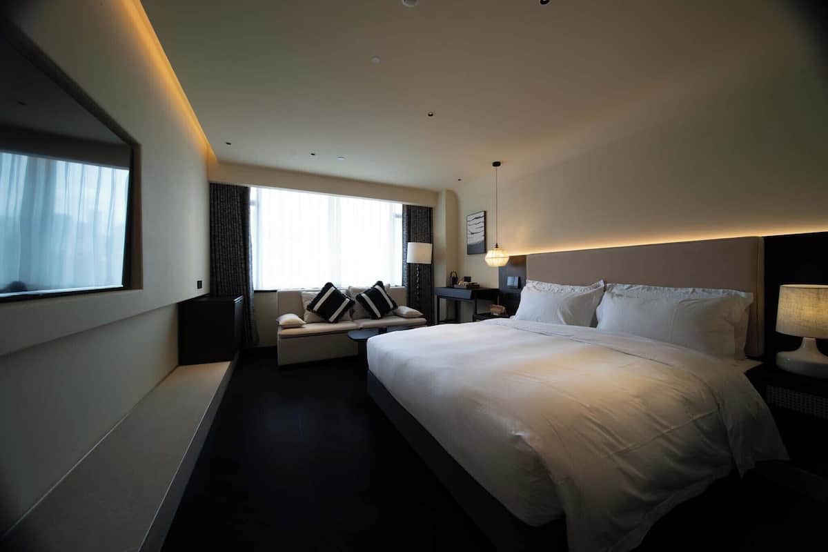 A modern hotel room with a large bed, a sofa, a desk, and a window with curtains.