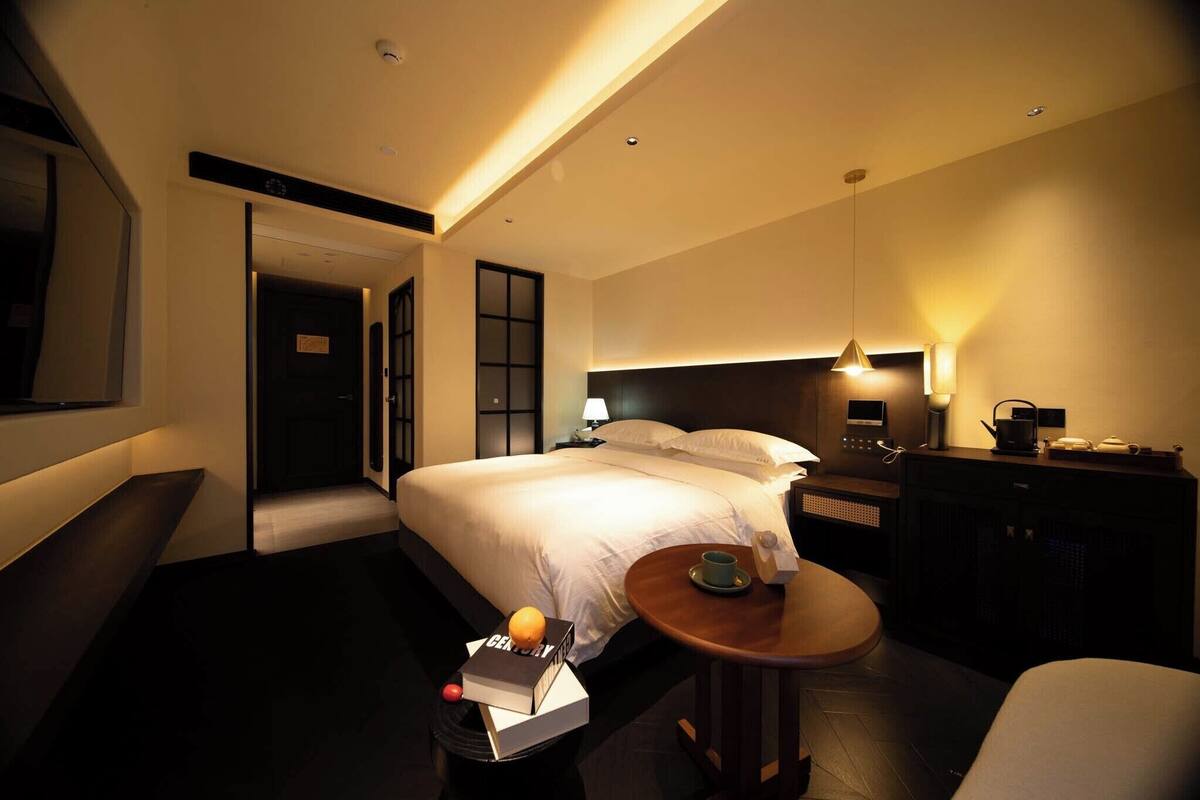 A hotel room with a bed, bedside table, lamp, and a small table with a cup and a book.