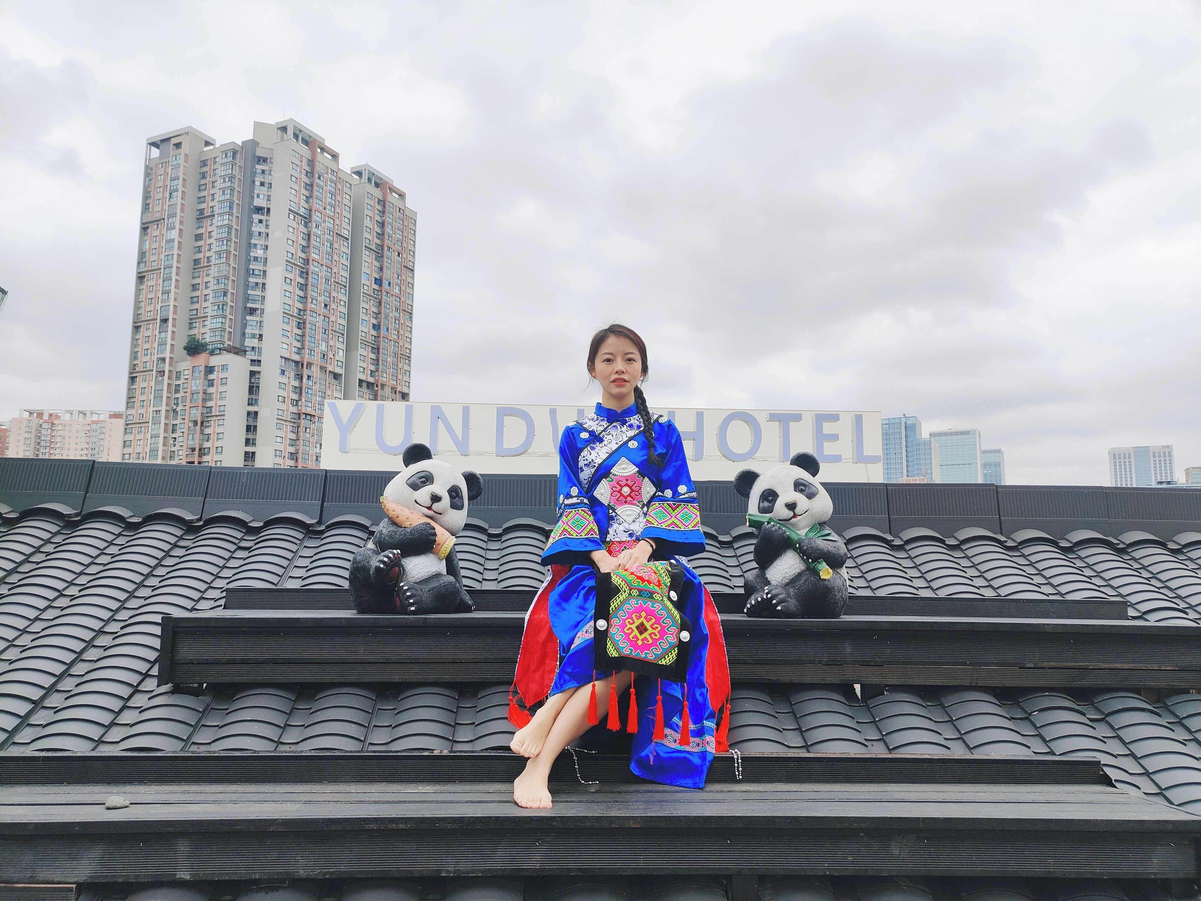 Une personne vêtue d’un costume traditionnel, assise sur un toit recouvert de tuiles, avec deux peluches de pandas.