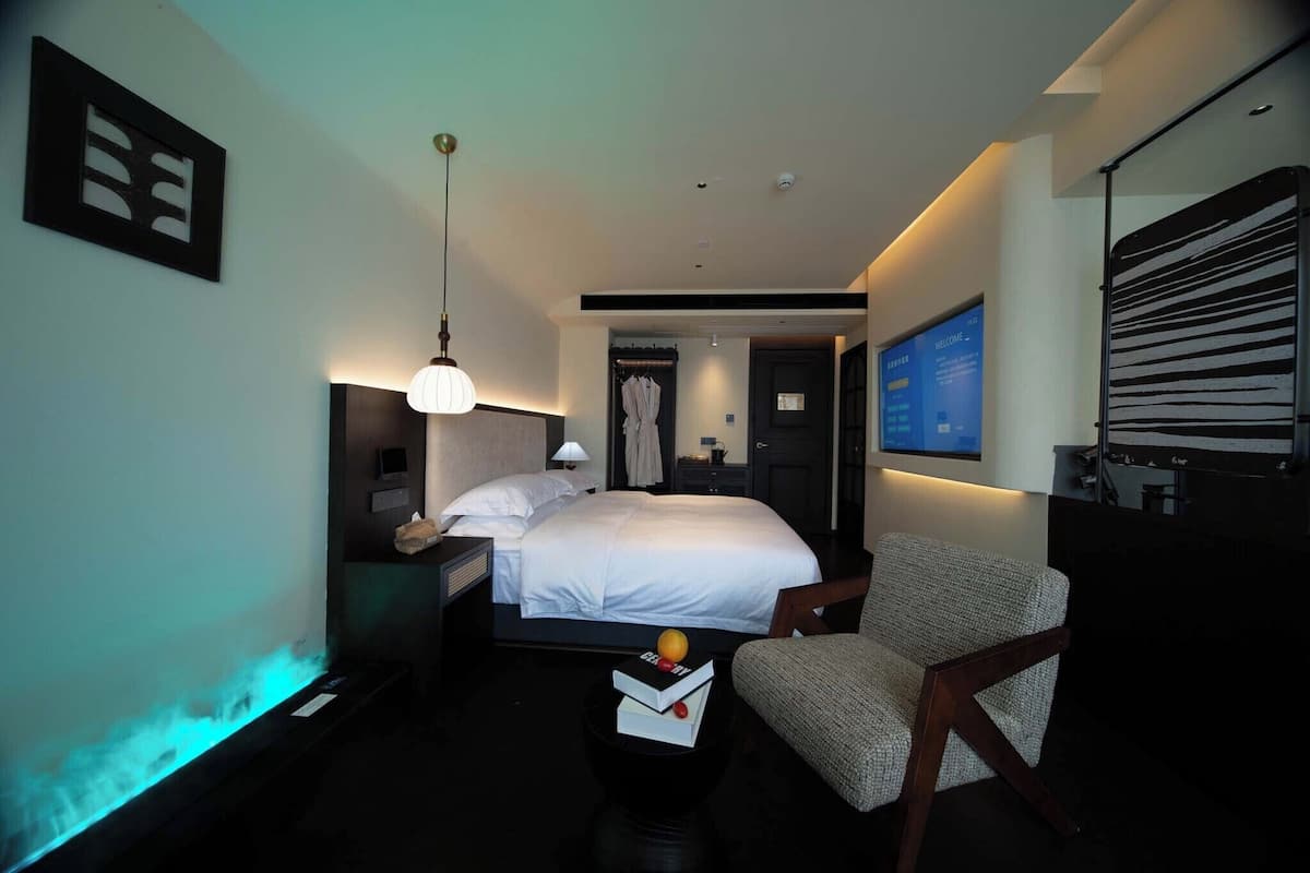 A modern hotel room with a large bed, bedside table, and a chair. There is a TV mounted on the wall and a window with a view.
