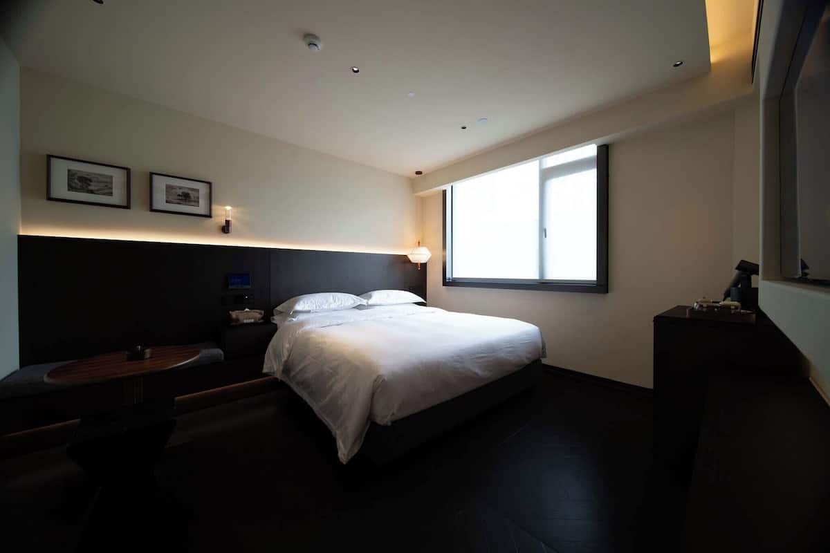 A modern hotel room with a large bed, a small table, and a window providing natural light.